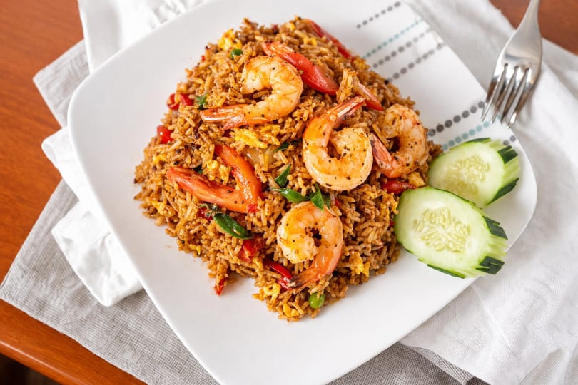 Shrimp fried rice with red peppers, green peas, and sliced cucumbers on a white plate.