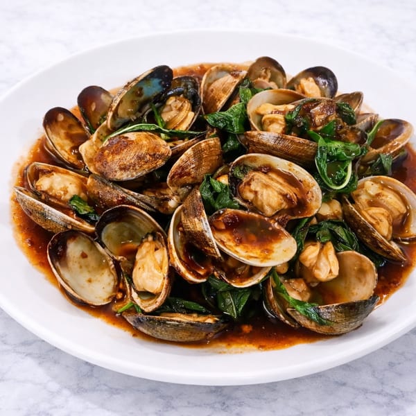 A white plate filled with stir-fried clams in a rich brown sauce with green basil leaves.