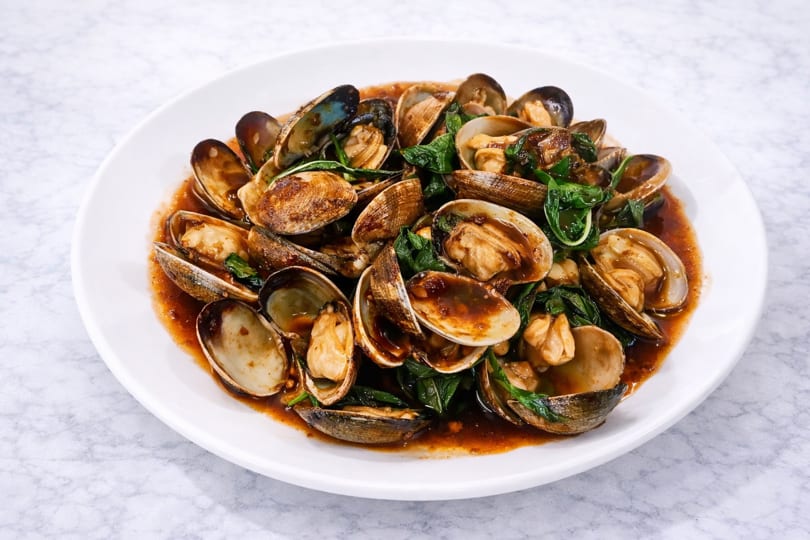 A white plate filled with stir-fried clams in a rich brown sauce with green basil leaves.